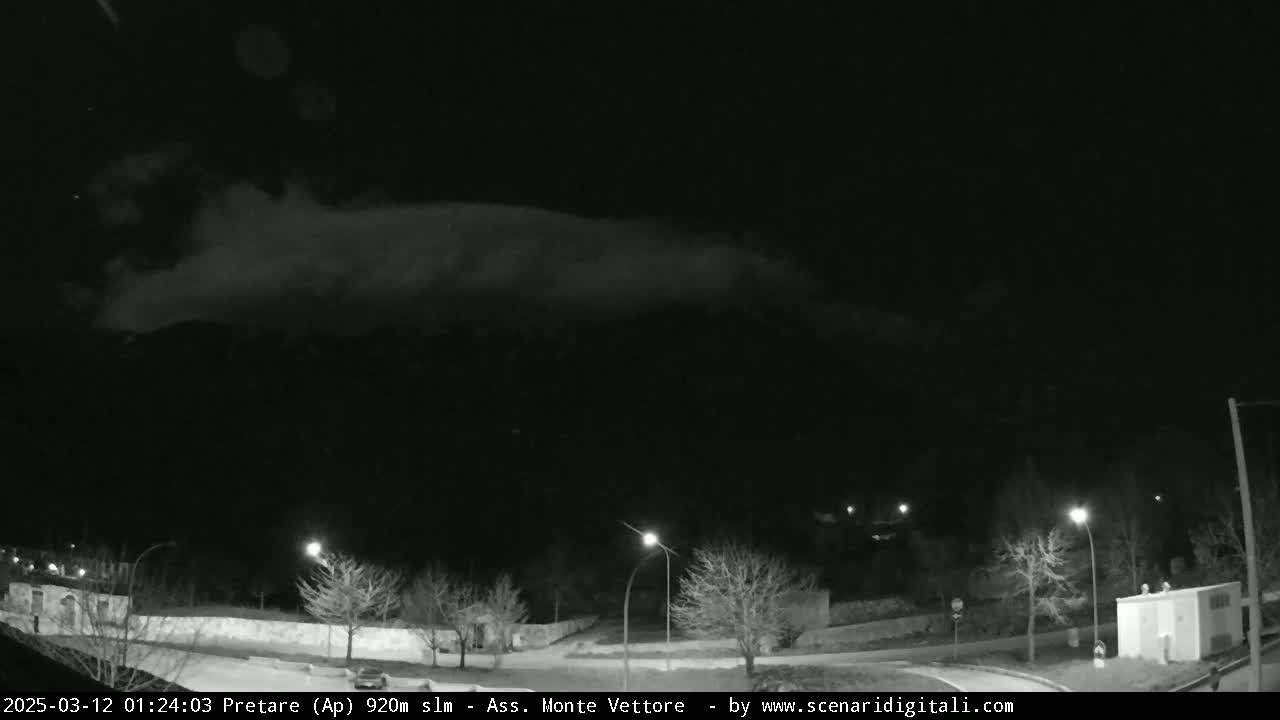A nighttime, low-light view shows a mostly clear sky with a low cloud bank over a dark mountain range behind a sparsely lit town.