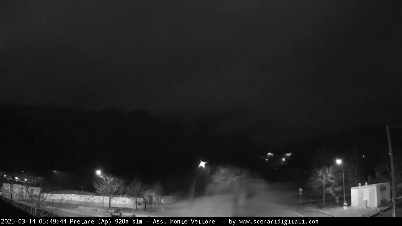 A nighttime, snowy scene shows a road curving through a sparsely lit area with trees and buildings under a dark sky.
