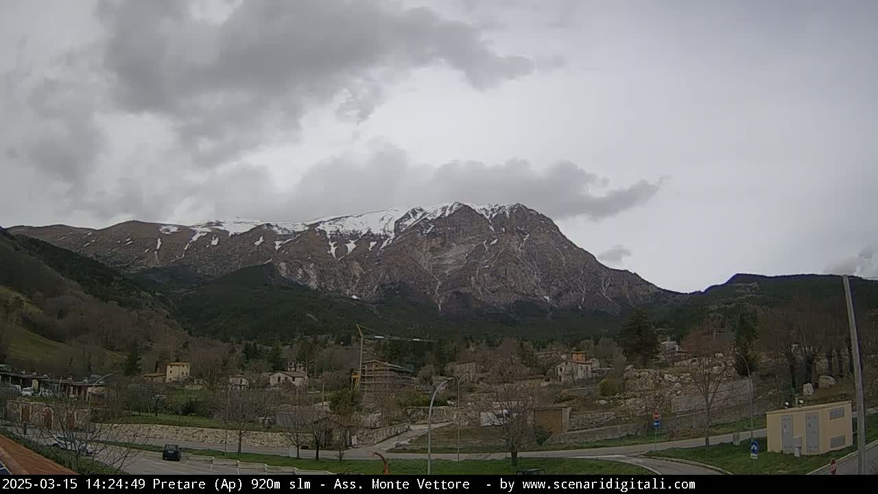 A snow-capped mountain looms over a small village nestled in a valley under a cloudy sky.