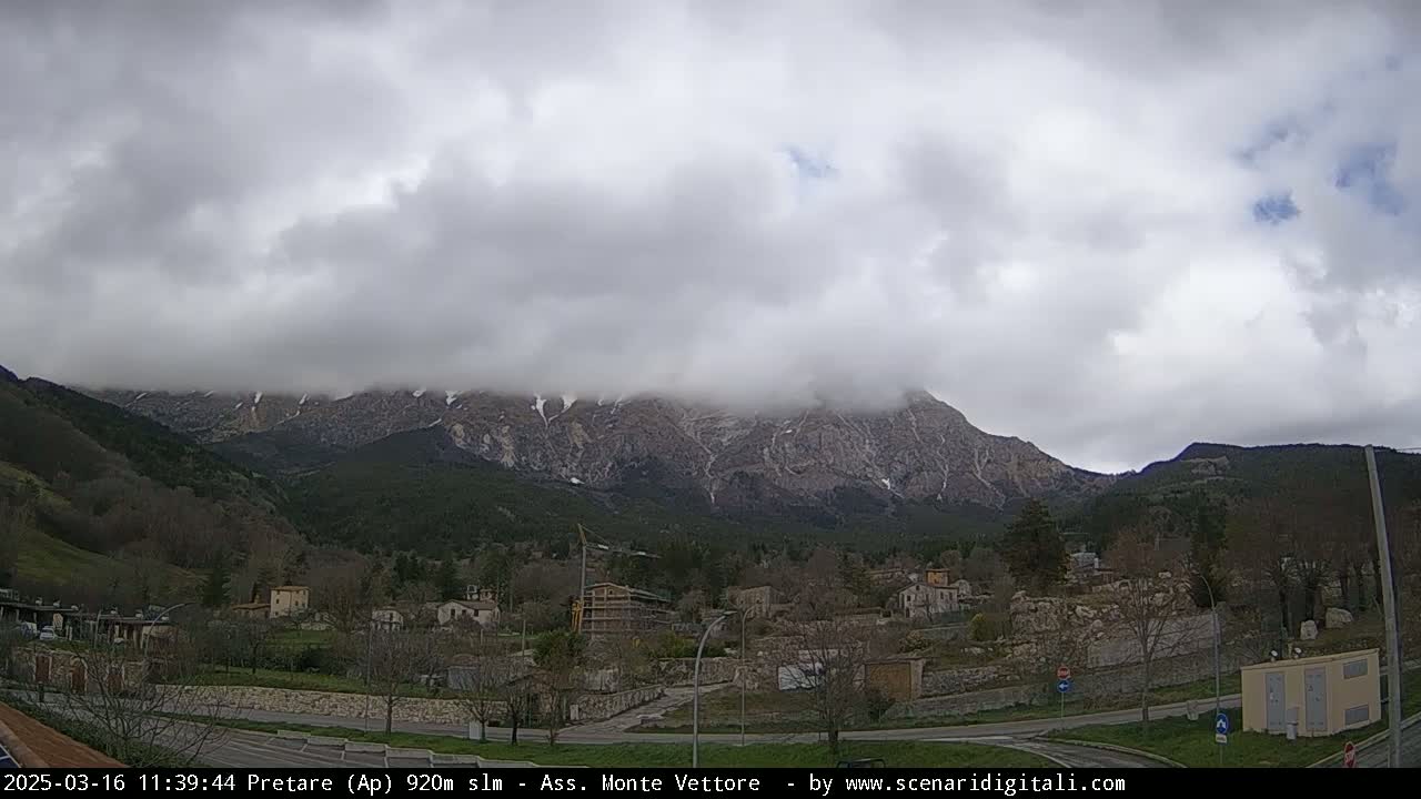 A small village nestled in a valley, with snow-capped mountains partially obscured by clouds in the background.