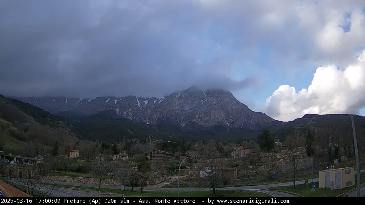 A partly cloudy sky overlooks a valley town nestled at the base of a large, snow-capped mountain partially obscured by clouds.