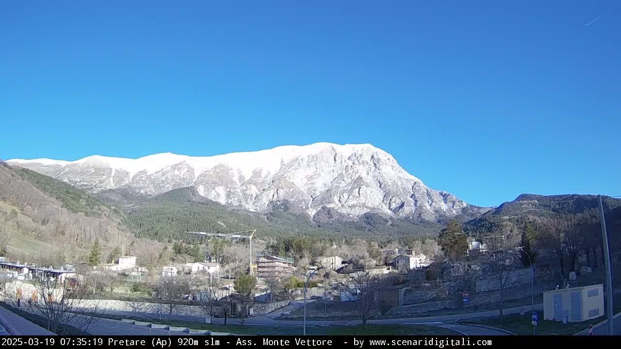 A snow-capped mountain looms over a small village nestled in a valley on a clear, sunny day.