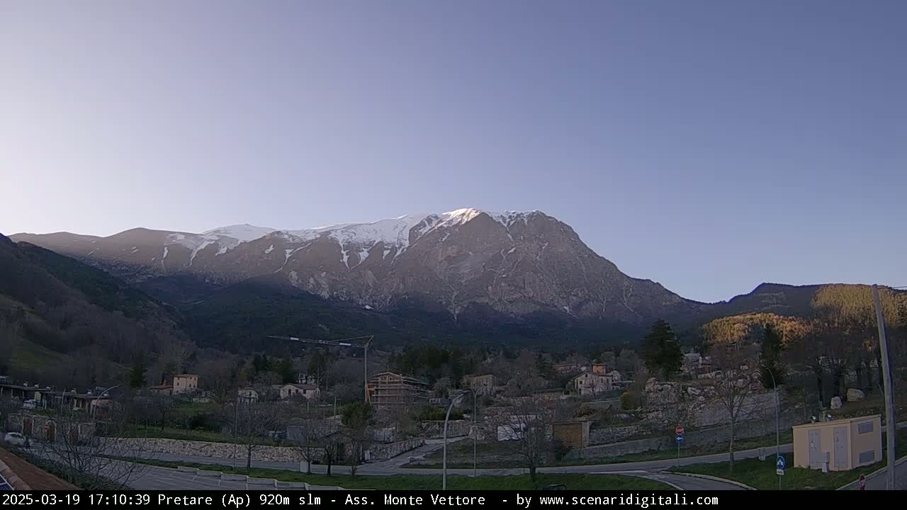 A snow-capped mountain rises behind a small town nestled in a valley under a clear, light-blue sky.