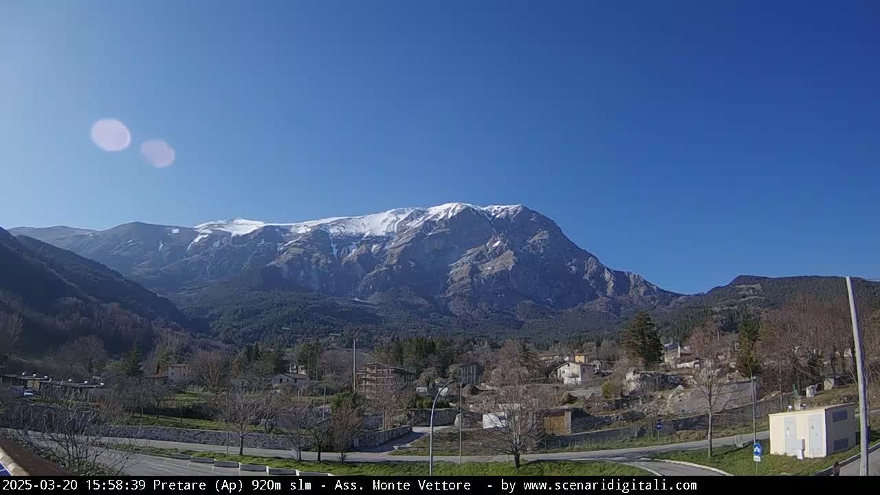 A snow-capped mountain looms over a small village nestled in a valley on a clear, sunny day.