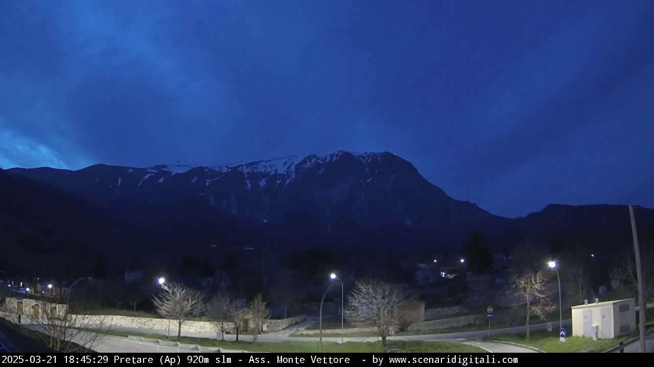 A snow-capped mountain looms over a small town at dusk under a clear, dark blue sky.