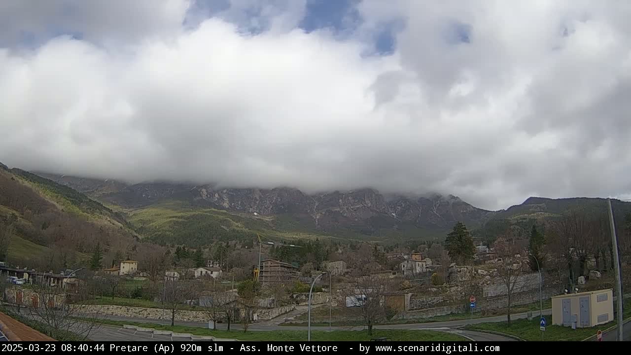 A small village nestled in a valley, with overcast skies and mountains shrouded in low clouds in the background.