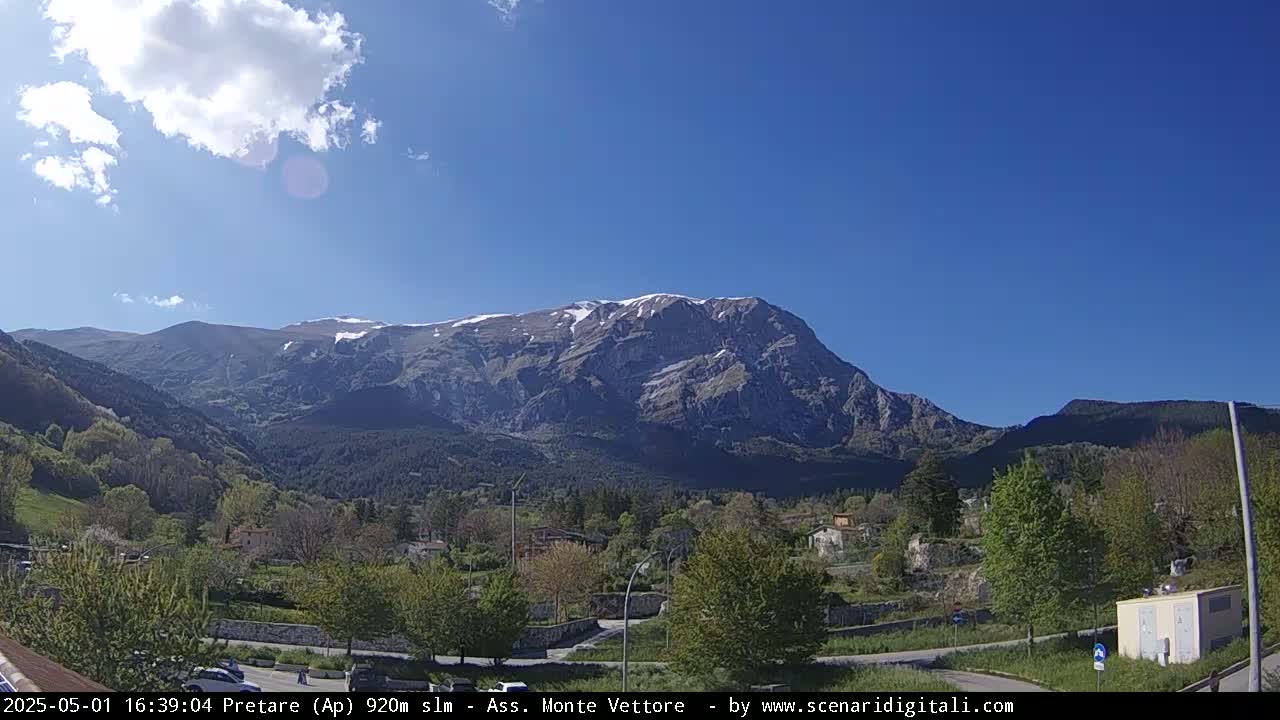 A snow-capped mountain rises in the background under a mostly sunny sky, overlooking a valley with a small town and lush green vegetation.