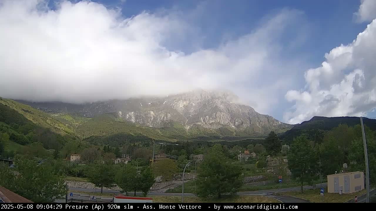 A partly cloudy sky hangs over a valley town nestled at the foot of a large, partially cloud-covered mountain range.