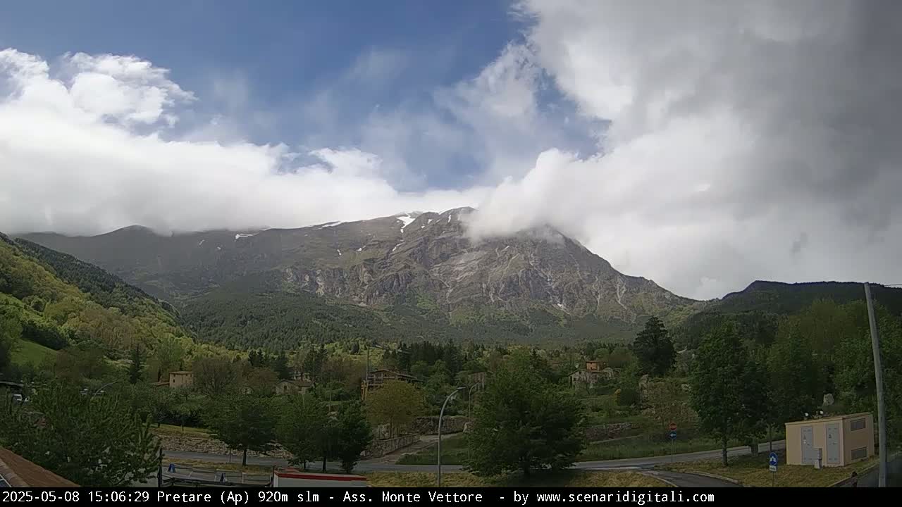 A partly cloudy day reveals a mountain range with some snow near its peak, partially obscured by clouds, overlooking a valley with green trees and buildings.