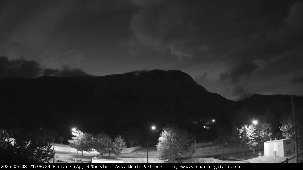 A nighttime, cloudy view of a mountain range silhouetted against the sky, with a few lights illuminating trees and a building in the foreground.