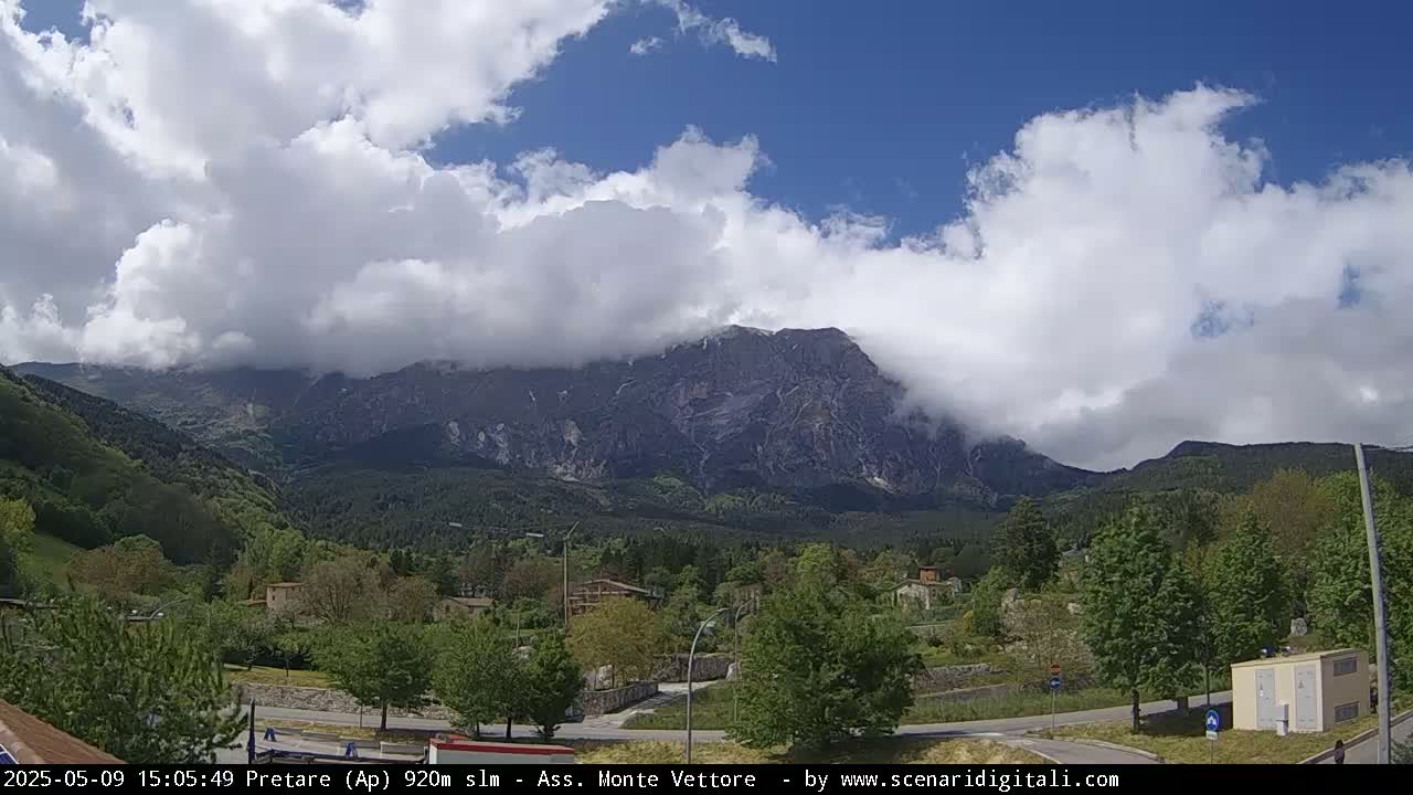 A partly cloudy day reveals a mountain range partially obscured by clouds, with a valley of green trees and buildings in the foreground.
