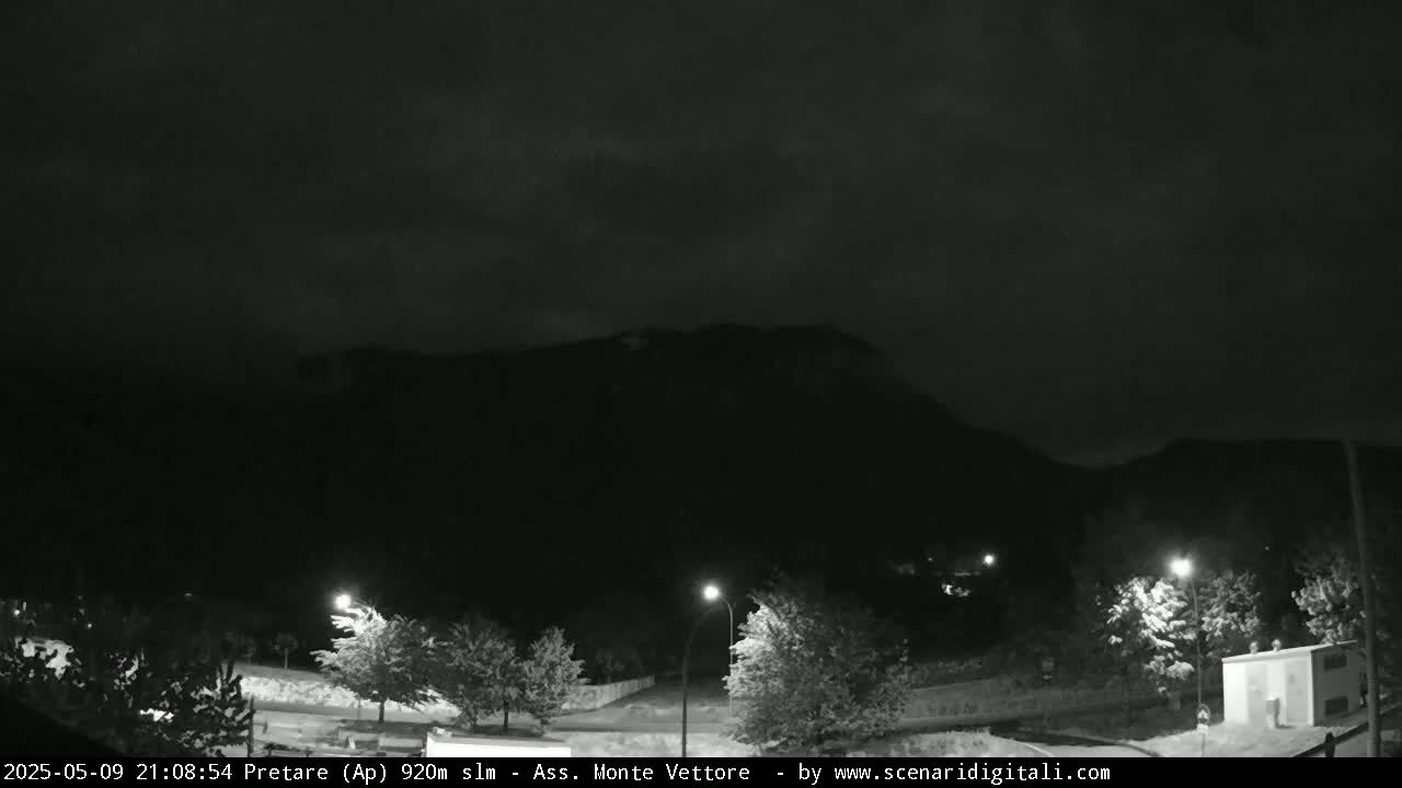 A nighttime view shows a dark mountain silhouetted against an overcast sky, with a few streetlights illuminating trees and a small building in the foreground.