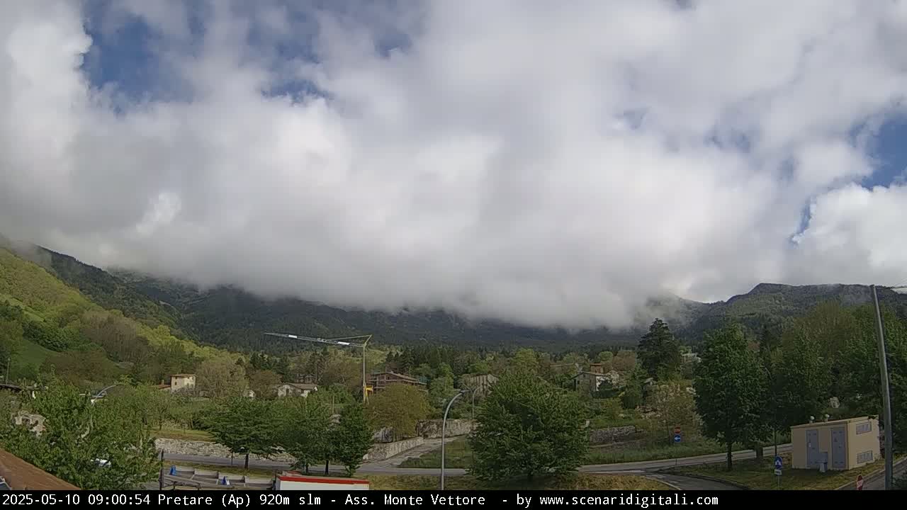 A cloudy, partly sunny day reveals a valley town nestled among green hills and mountains shrouded in fog.