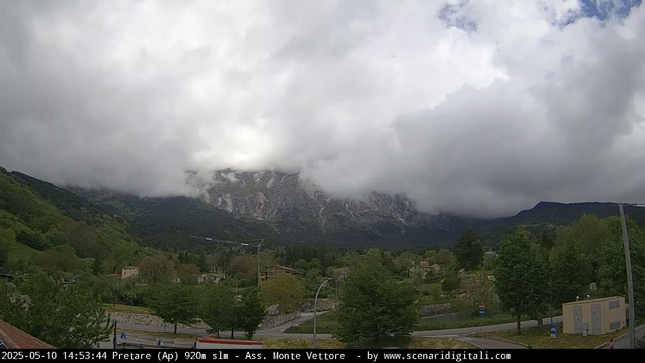 A cloudy mountain range is visible in the distance behind a valley of green trees and small buildings, under an overcast sky.
