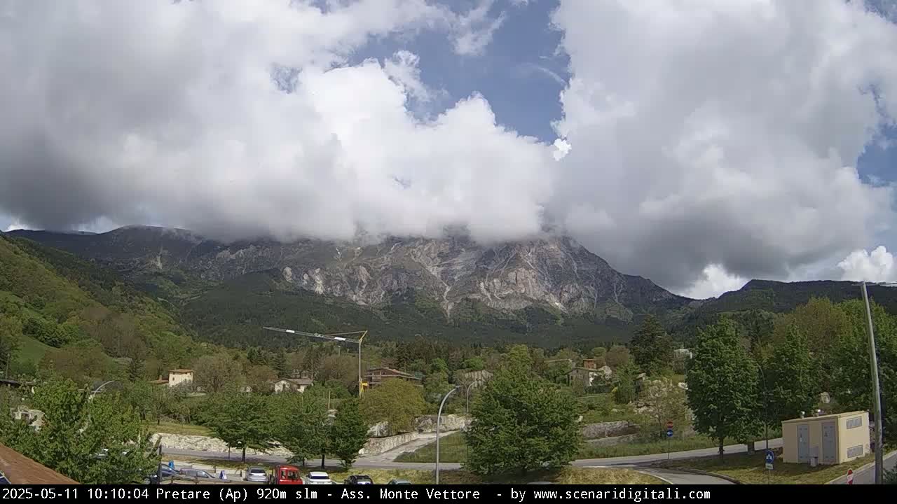 A partly cloudy sky hangs over a mountain range, partially obscured by clouds, with a town nestled in a valley at the base of the mountains.