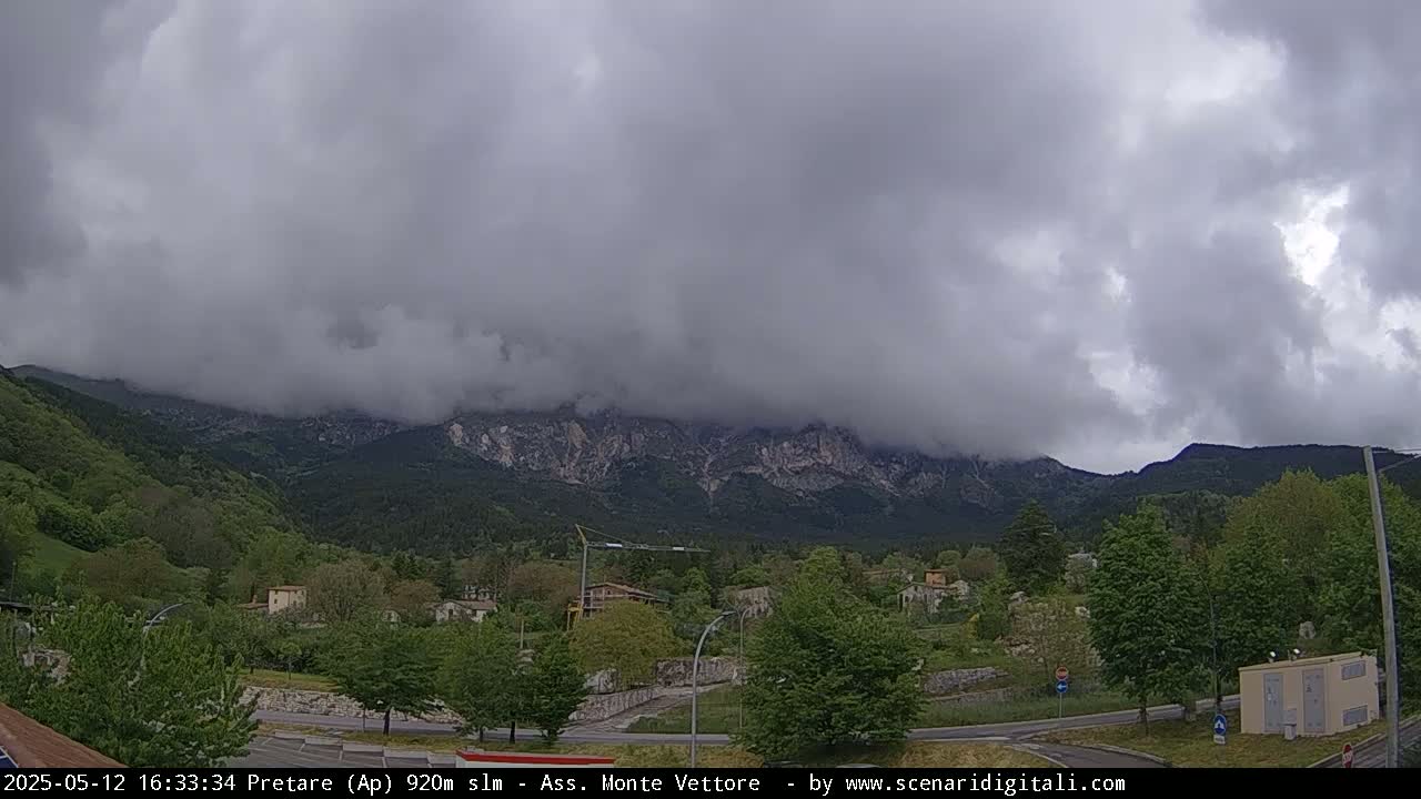 A cloudy day reveals a mountain range partially obscured by fog, overlooking a small town nestled in a valley of green trees and roads.
