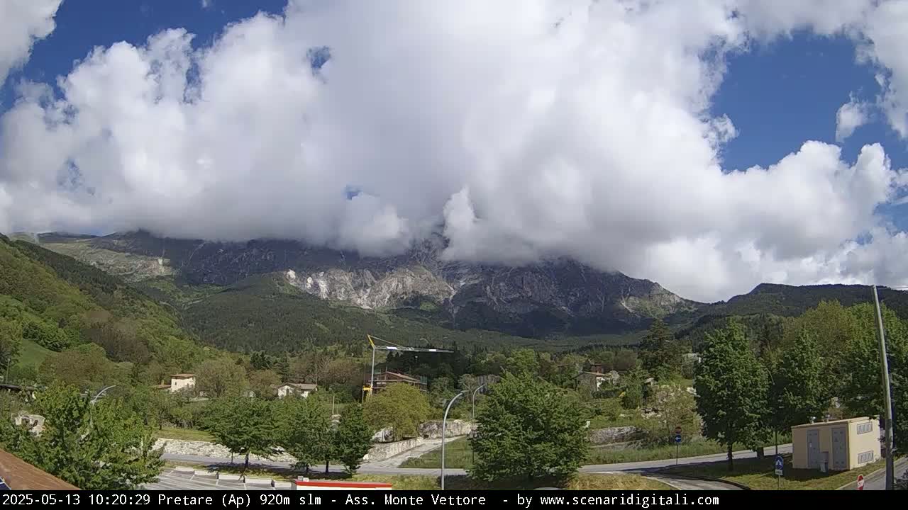 A partly cloudy day reveals a mountain range partially obscured by clouds, with green trees and a small town nestled in the valley below.