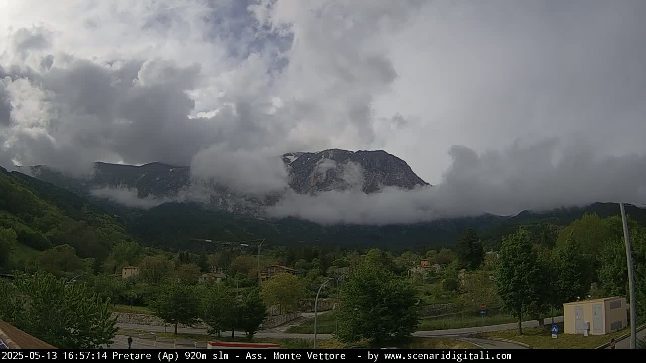 A mountain range partially obscured by clouds is visible from a valley with green trees and buildings, under an overcast sky.