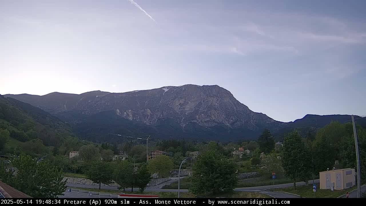 A large, rocky mountain with some snow near the peak is visible in the background, with a small town and green trees in the foreground under a mostly clear, light blue sky.