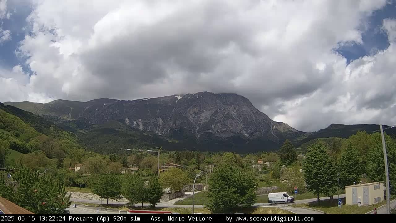 A large, rocky mountain with some snow near the peak is visible behind a town, under a partly cloudy sky.
