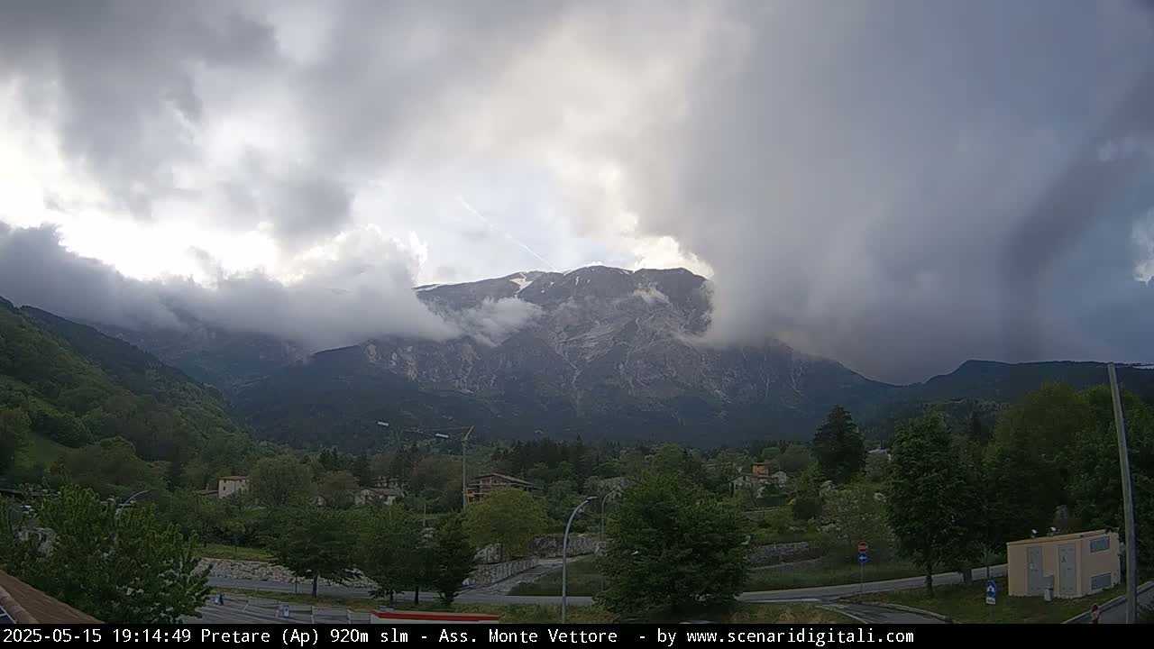 A mountain range partially obscured by clouds is visible in the distance beyond a valley of green trees and small buildings under an overcast sky.