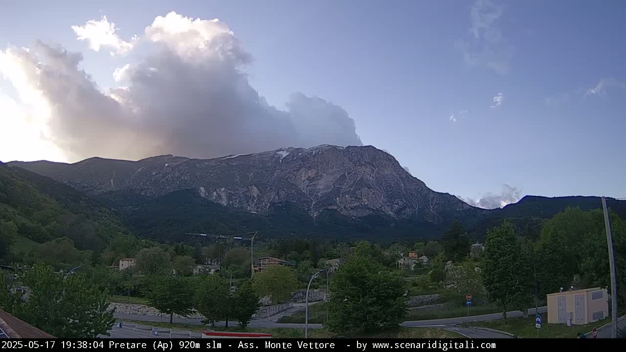 A large, rocky mountain with some snow near the peak is visible under a partly cloudy sky, behind a valley with green trees and a small town.