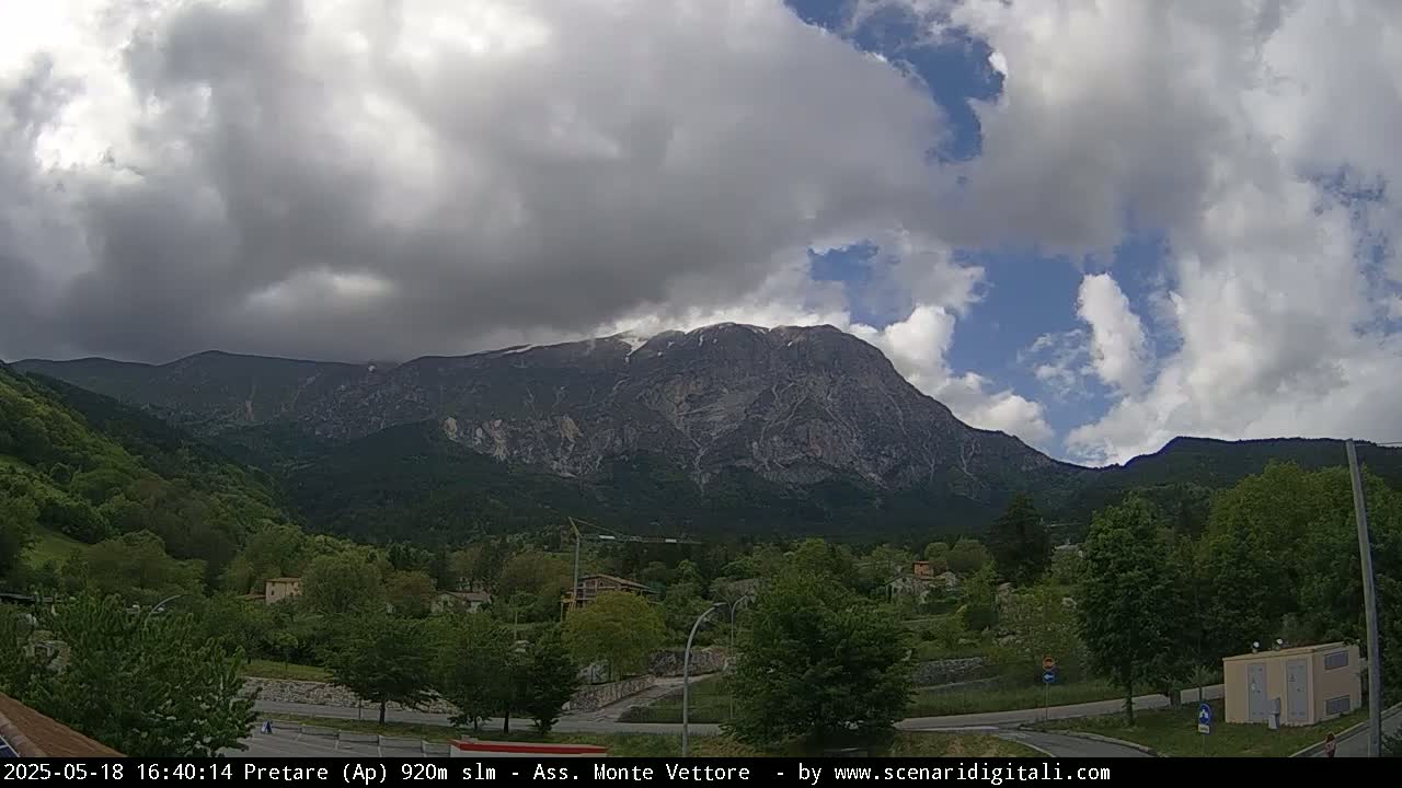 A partly cloudy sky overlooks a valley of green trees and small buildings, with a large, rocky mountain dominating the background.