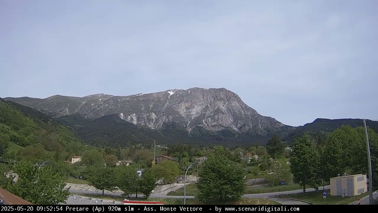 A large, rocky mountain with patches of snow sits in the background of a lush green valley, under a mostly clear sky.
