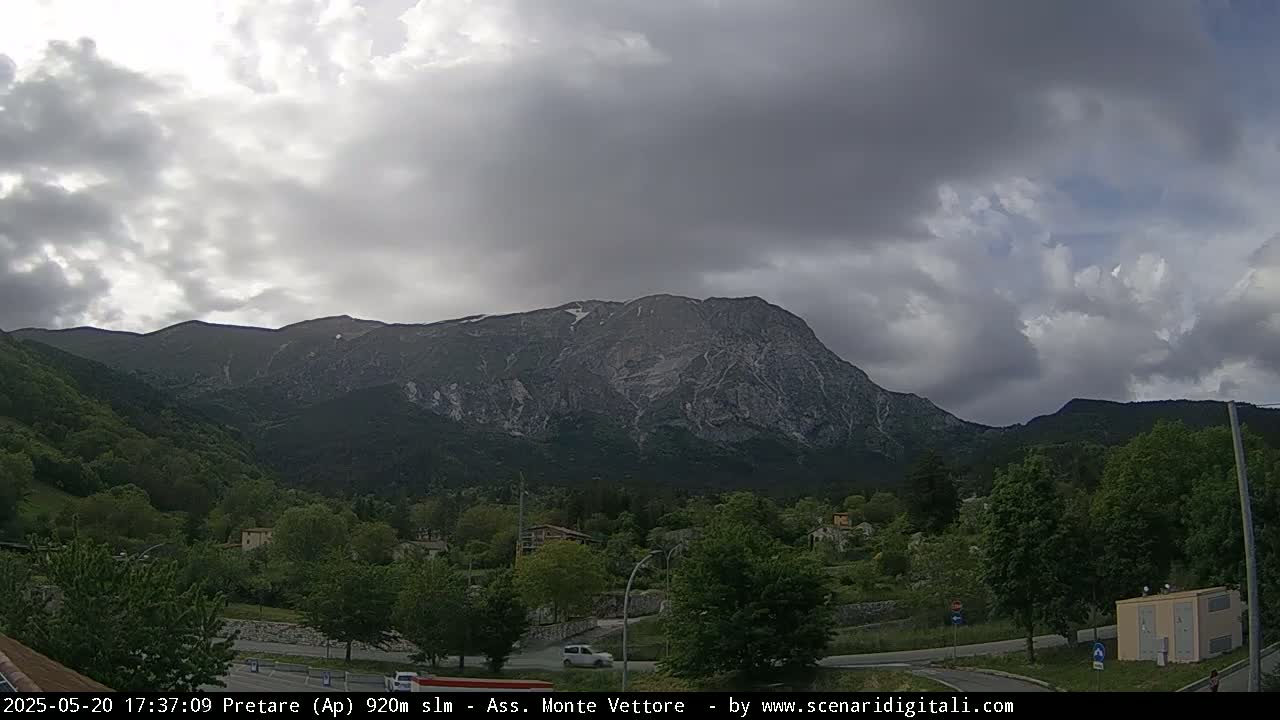 A large, rocky mountain with patches of snow is visible behind a valley of green trees and buildings under a mostly cloudy sky.