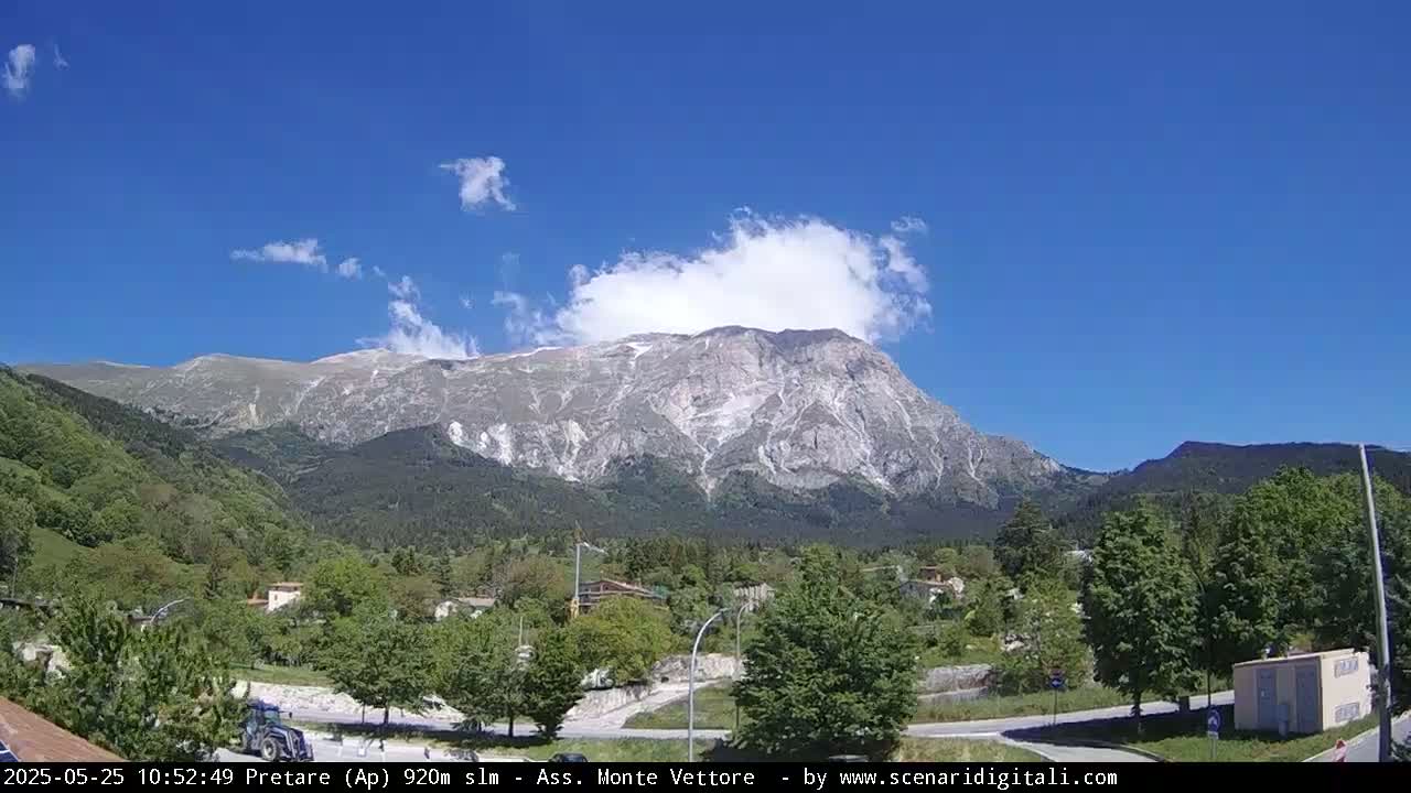 A large, rocky mountain with some snow near its peak is visible behind a valley of green trees and a small town under a mostly clear blue sky with a few fluffy clouds.