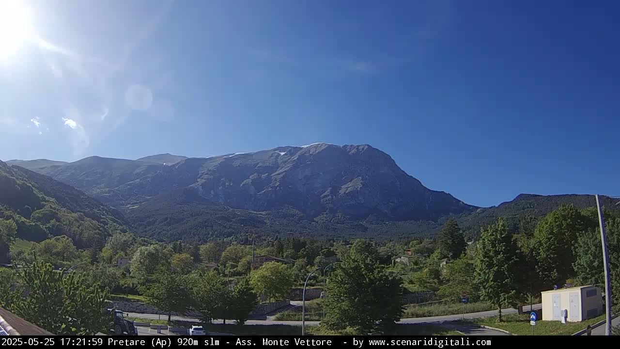 A sunlit mountain range with a snow-capped peak overlooks a verdant valley and small village under a clear blue sky.