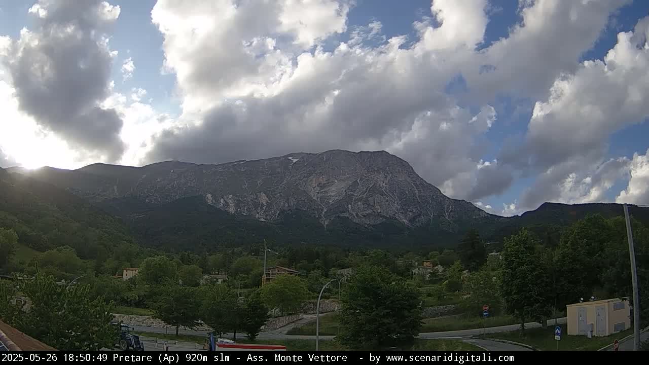 A partly cloudy sky looms over a mountain range, with a valley of green trees and buildings in the foreground.