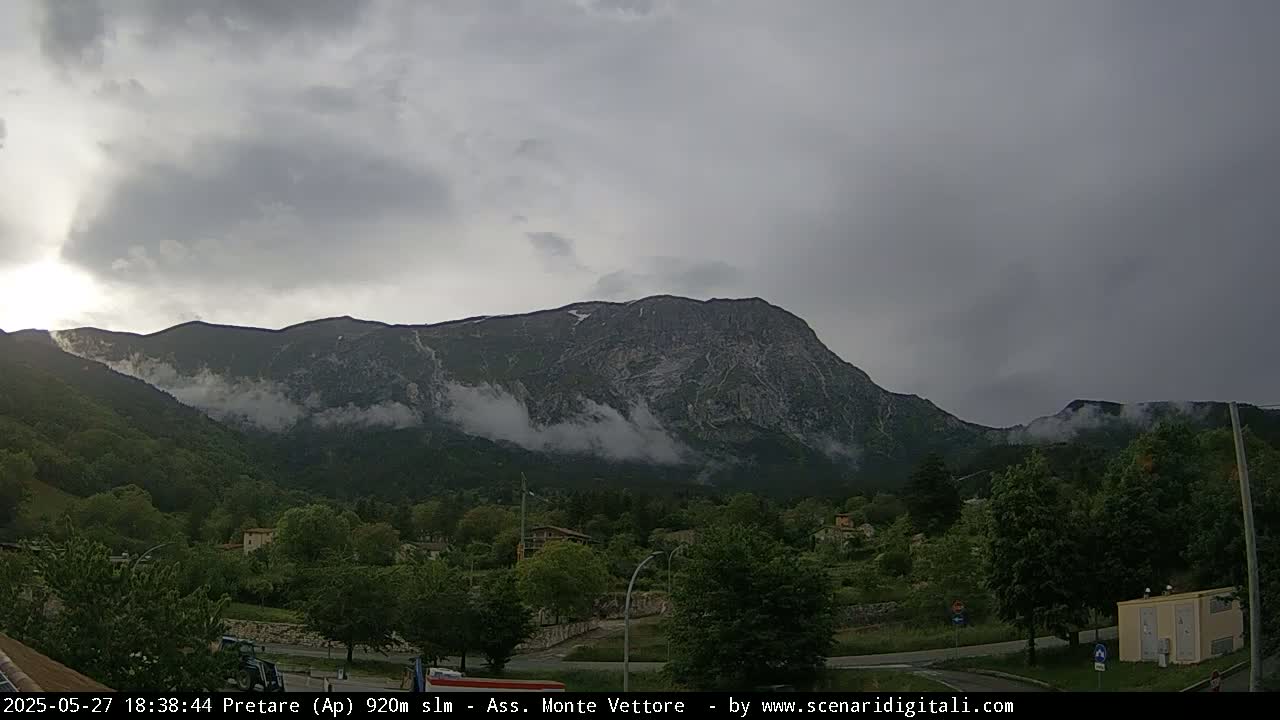 A large, rocky mountain range partially shrouded in fog is visible under an overcast sky, with green vegetation and a few buildings in the foreground.