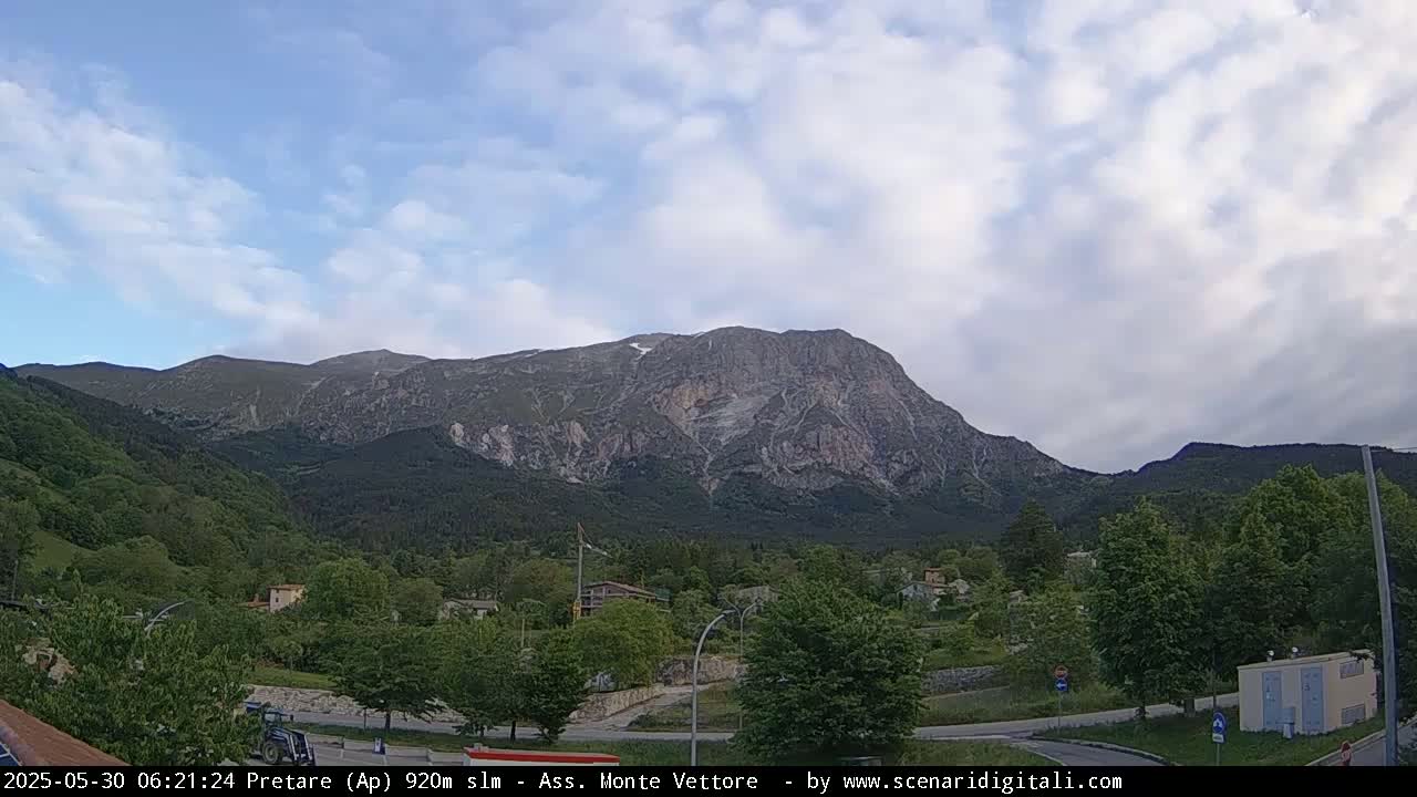 A large, rocky mountain dominates the view, under a partly cloudy sky, overlooking a valley with lush green trees and a small town.