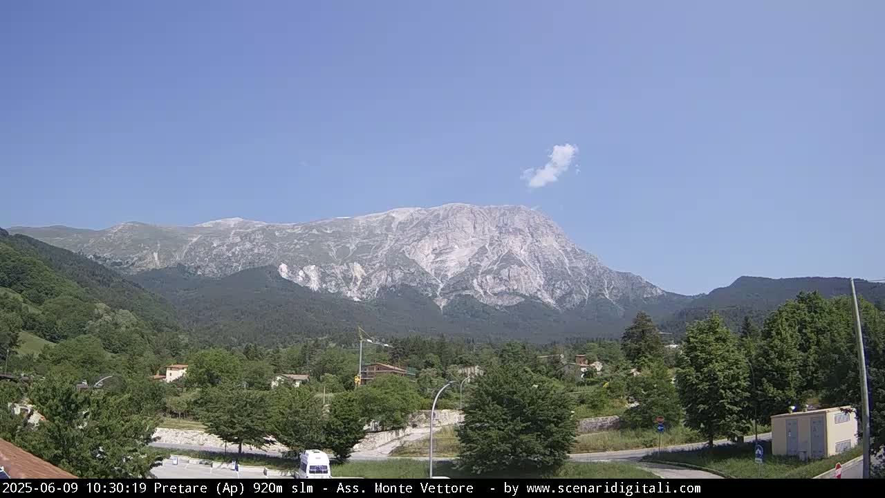 A large, light-grey mountain with a few patches of snow is visible in the distance, under a mostly clear blue sky with a small, wispy cloud, overlooking a valley of green trees and a few buildings.