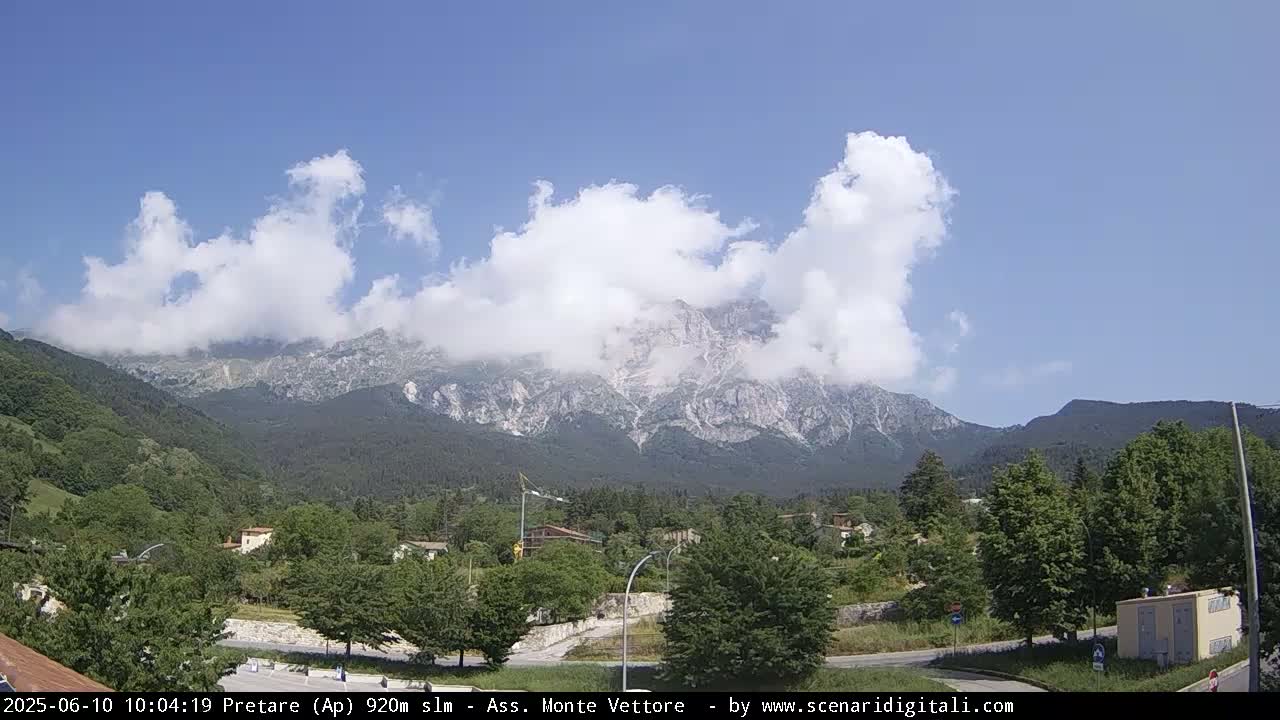 A partly cloudy day reveals a mountain range behind a green valley with a town nestled amongst the trees.