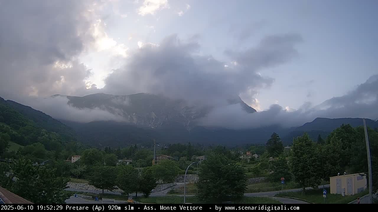 A partly cloudy sky hangs over a valley with green trees and a large mountain partially obscured by fog.
