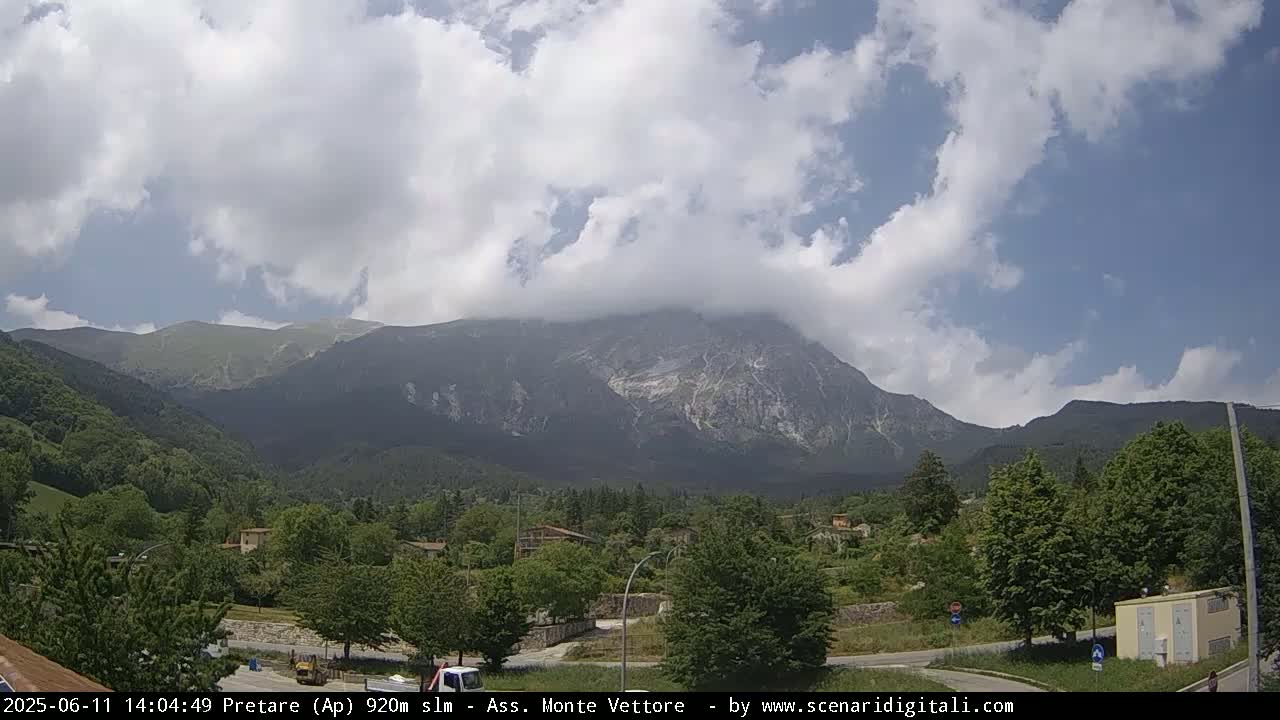 A partly cloudy day reveals a mountain range partially obscured by clouds, with a valley of green trees and buildings in the foreground.
