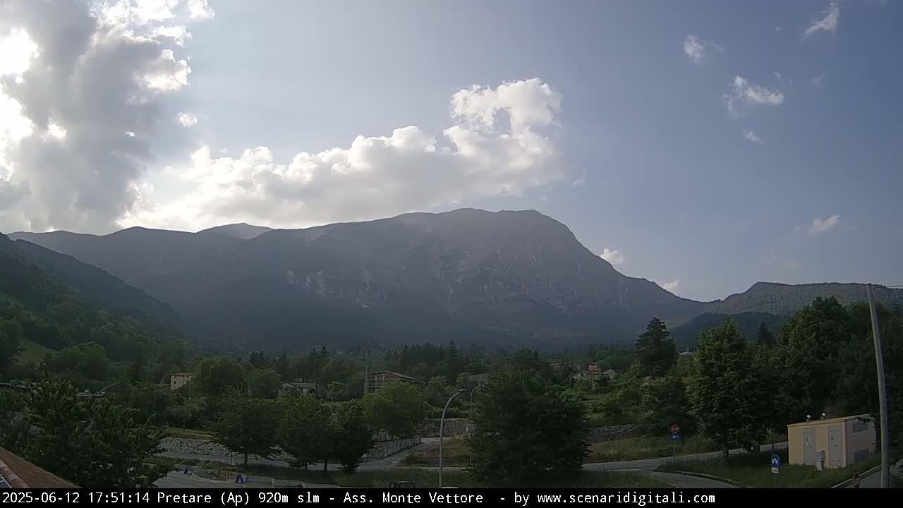 A partly cloudy sky overlooks a valley of green trees and buildings, with a large mountain range in the background.