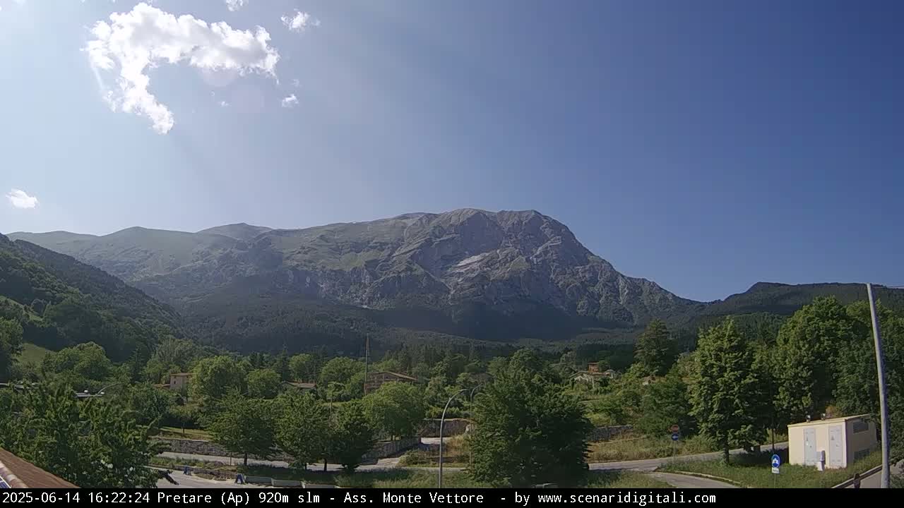 A large, rocky mountain dominates the background under a mostly sunny sky, overlooking a village nestled in a valley of green trees.