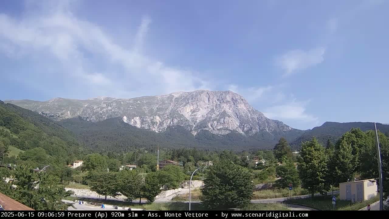 A large, light-colored mountain range is visible in the distance under a mostly clear, bright blue sky with a few wispy clouds, overlooking a valley with green trees and a small town.