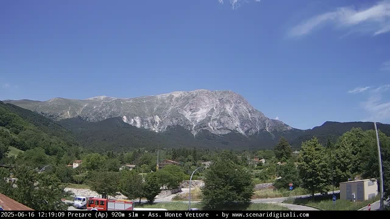 A large, light-grey mountain dominates the background under a mostly clear, blue sky with a few scattered clouds, overlooking a small town nestled amongst green trees and vegetation.
