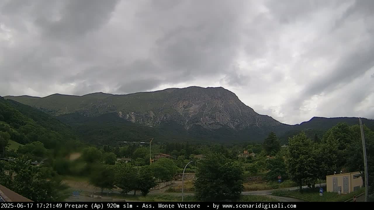 A large, rocky mountain dominates the landscape under a cloudy sky, with green trees and a small town nestled at its base.