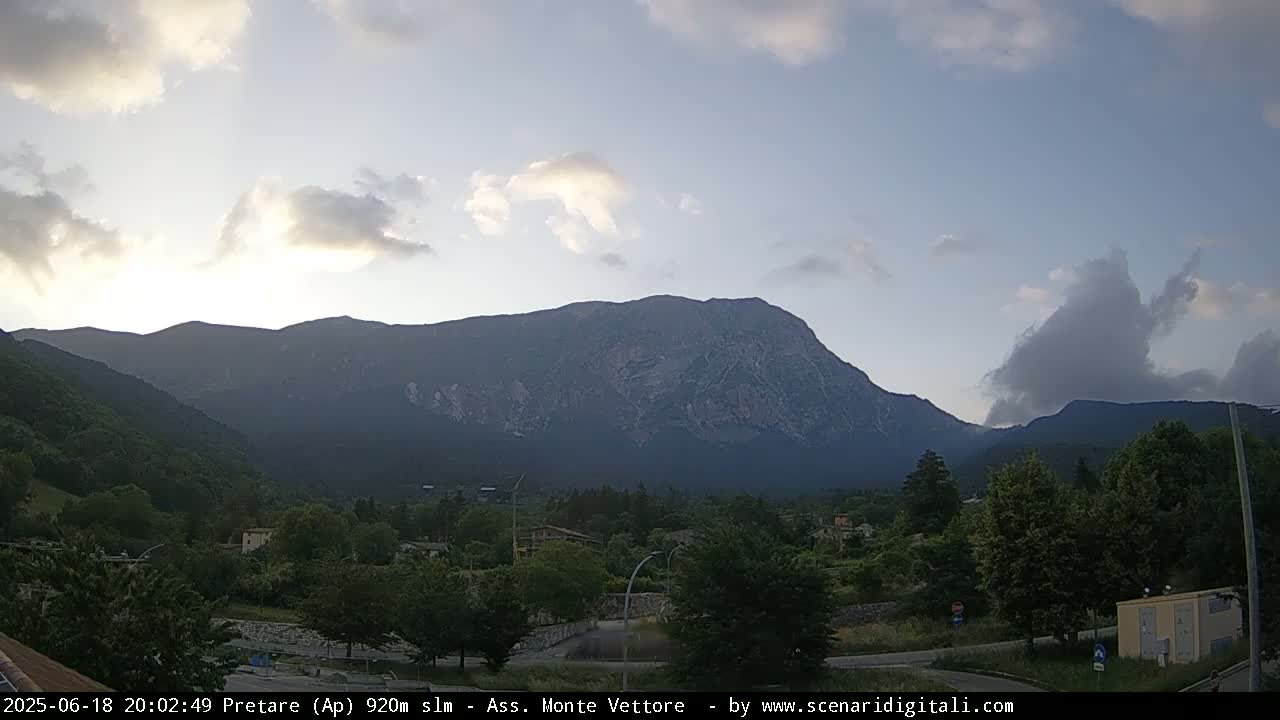 A partly cloudy evening sky overlooks a mountain range and a village nestled in a valley below.