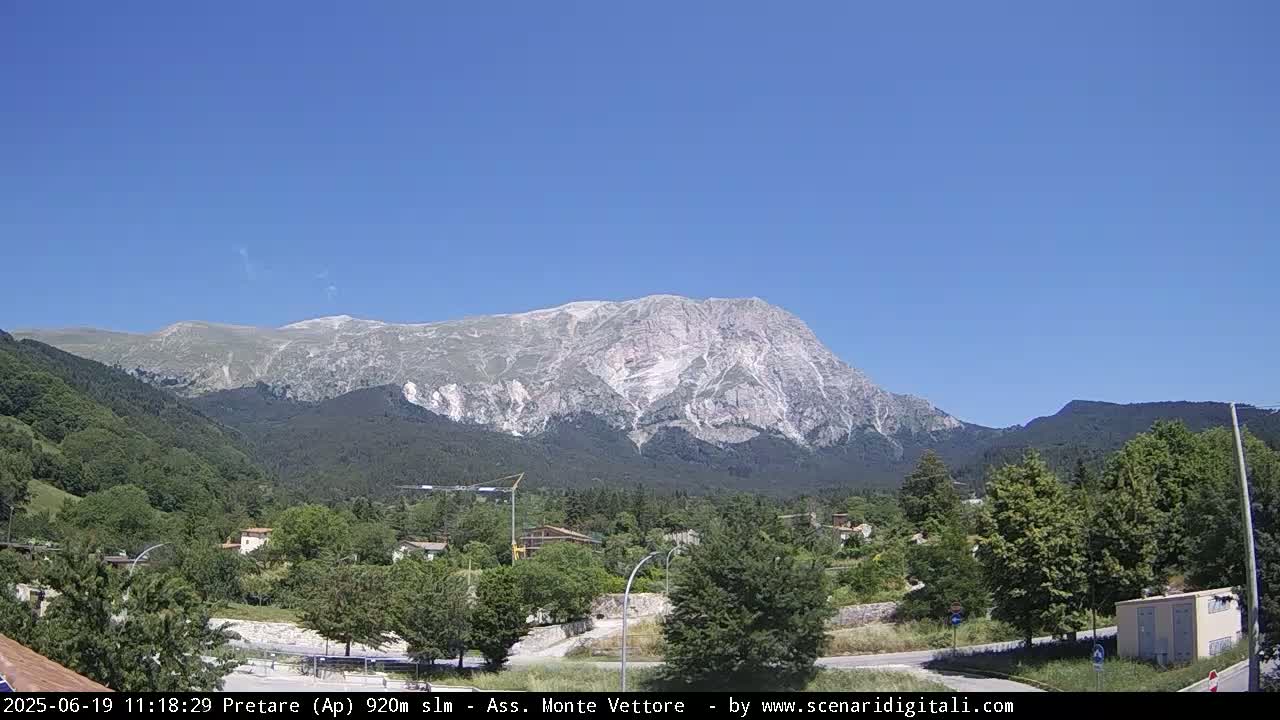 A large, light-grey mountain dominates the background under a clear blue sky, overlooking a valley of green trees and small buildings.