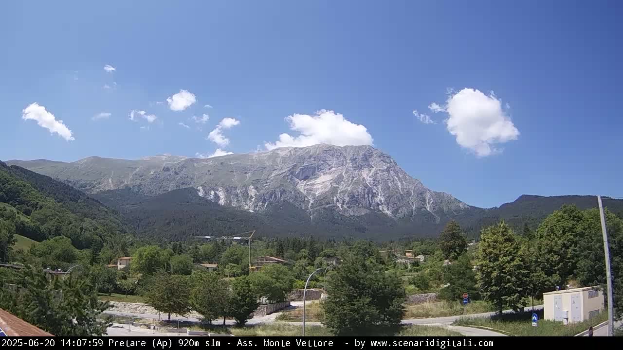 A large, rocky mountain dominates the background under a mostly sunny sky with scattered cumulus clouds, overlooking a valley of green trees and buildings.
