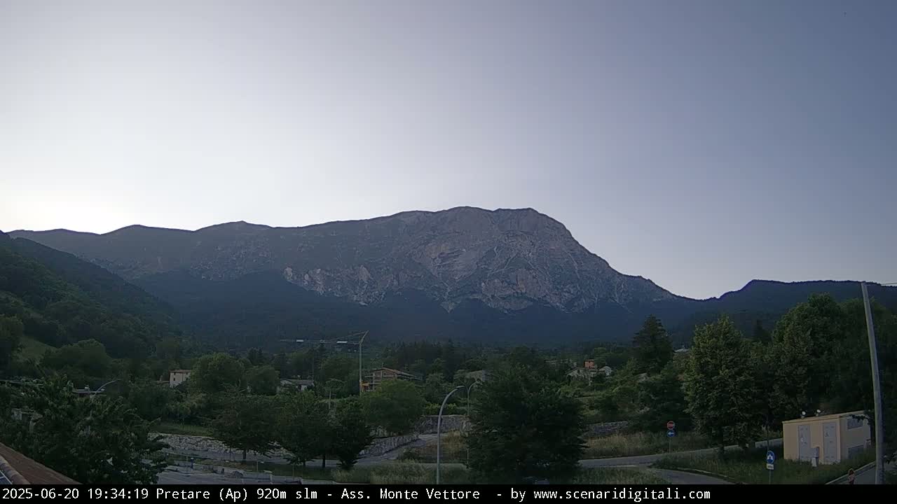 A large, light-grey mountain range dominates the background under a pale, clear sky, overlooking a valley of green trees and a small town.