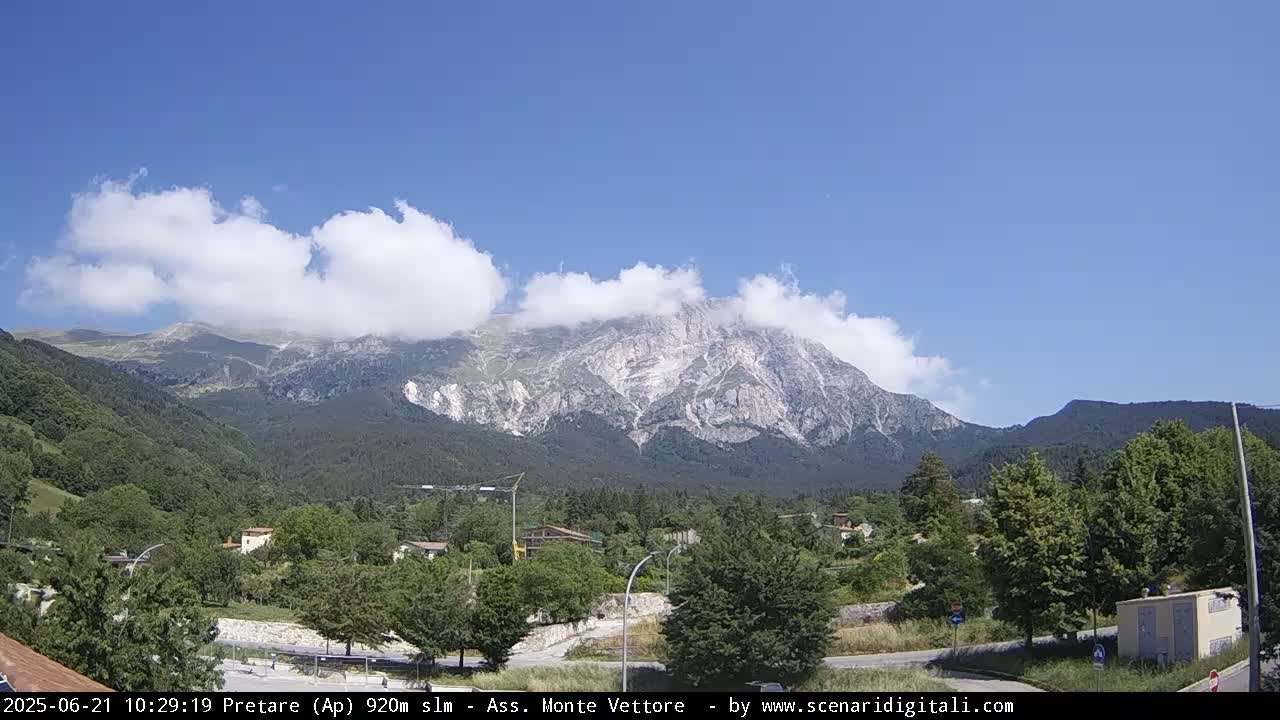 A partly cloudy day reveals a mountain range partially obscured by clouds, with a valley of green trees and buildings in the foreground.