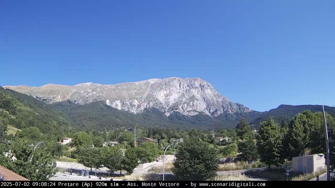A large, light-grey mountain dominates the background under a clear blue sky, overlooking a valley of green trees and a small town.