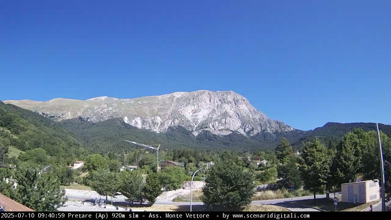 A large, light-colored mountain rises in the background under a clear blue sky, overlooking a valley of green trees and a small town.